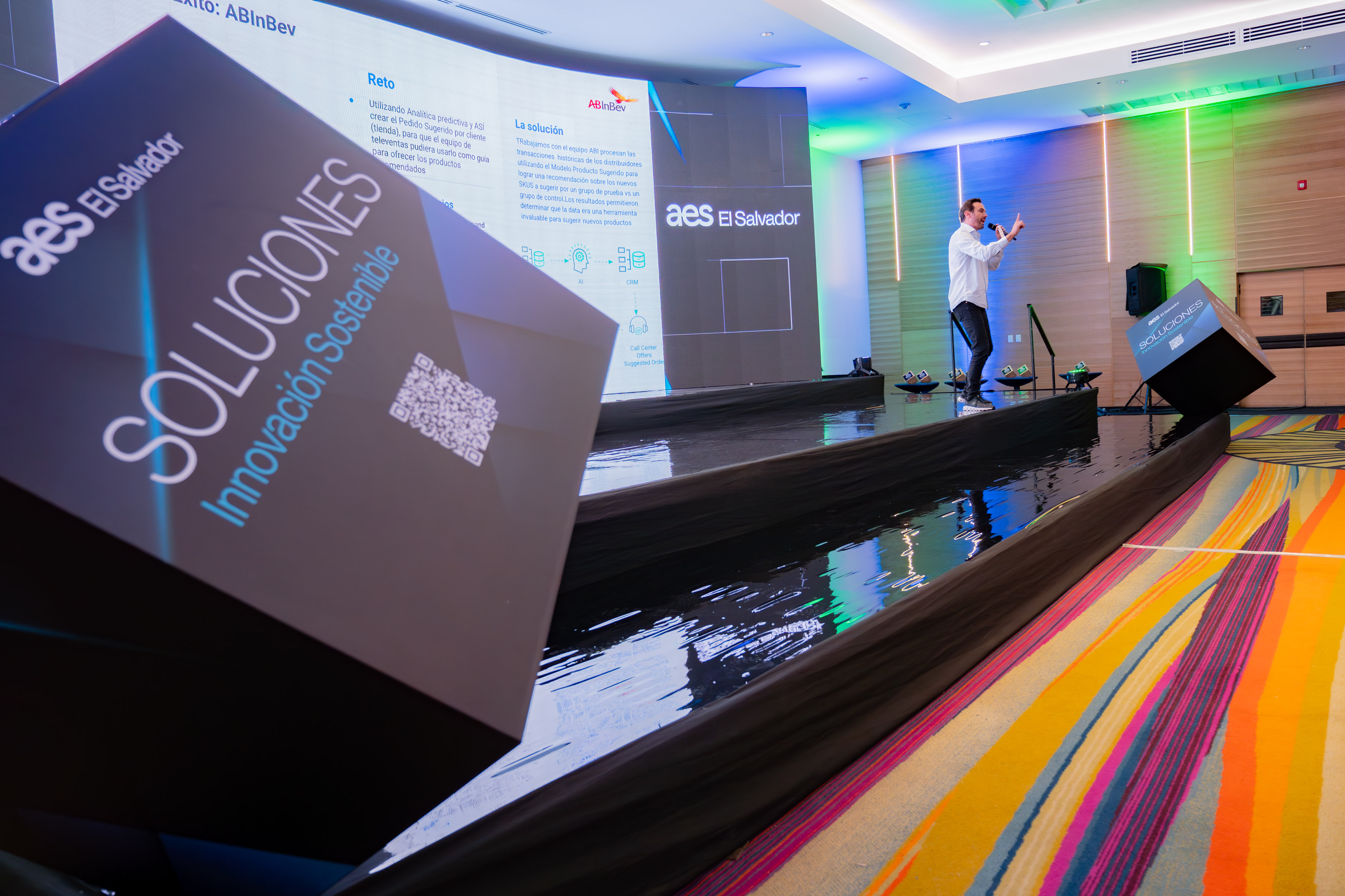 A presentation event by AES El Salvador featuring a speaker on stage discussing innovative sustainable solutions. The stage is surrounded by reflective black surfaces, with vibrant lighting and colorful carpet in the room. A large display screen shows project details, while a branded cube in the foreground reads "Soluciones Innovación Sostenible" with a QR code.