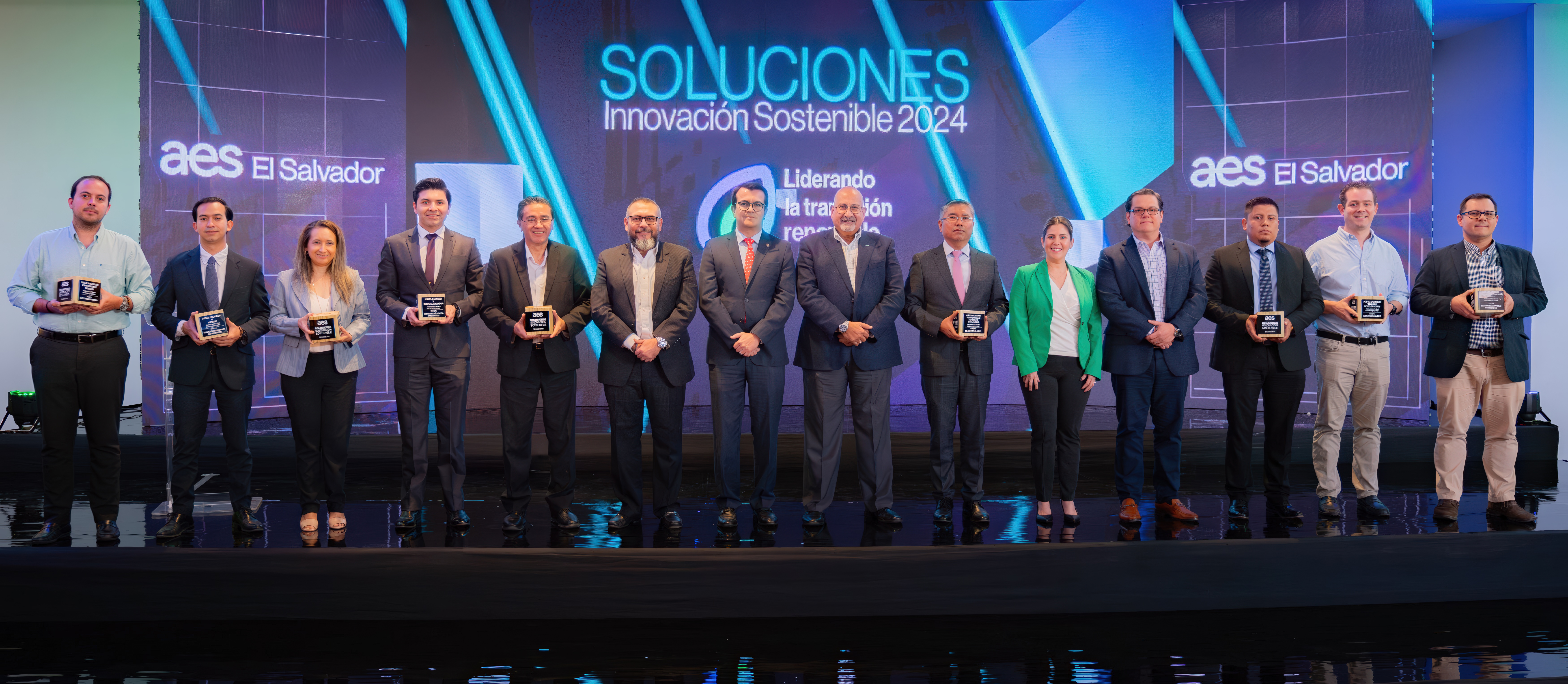 A group of 14 individuals standing on stage at the AES El Salvador Sustainable Solutions 2024 event. Each person is holding an award plaque, symbolizing recognition for their contributions. The background features a vibrant event banner with the words "Soluciones Innovación Sostenible 2024." The group is dressed in business attire, highlighting a formal and celebratory occasion.