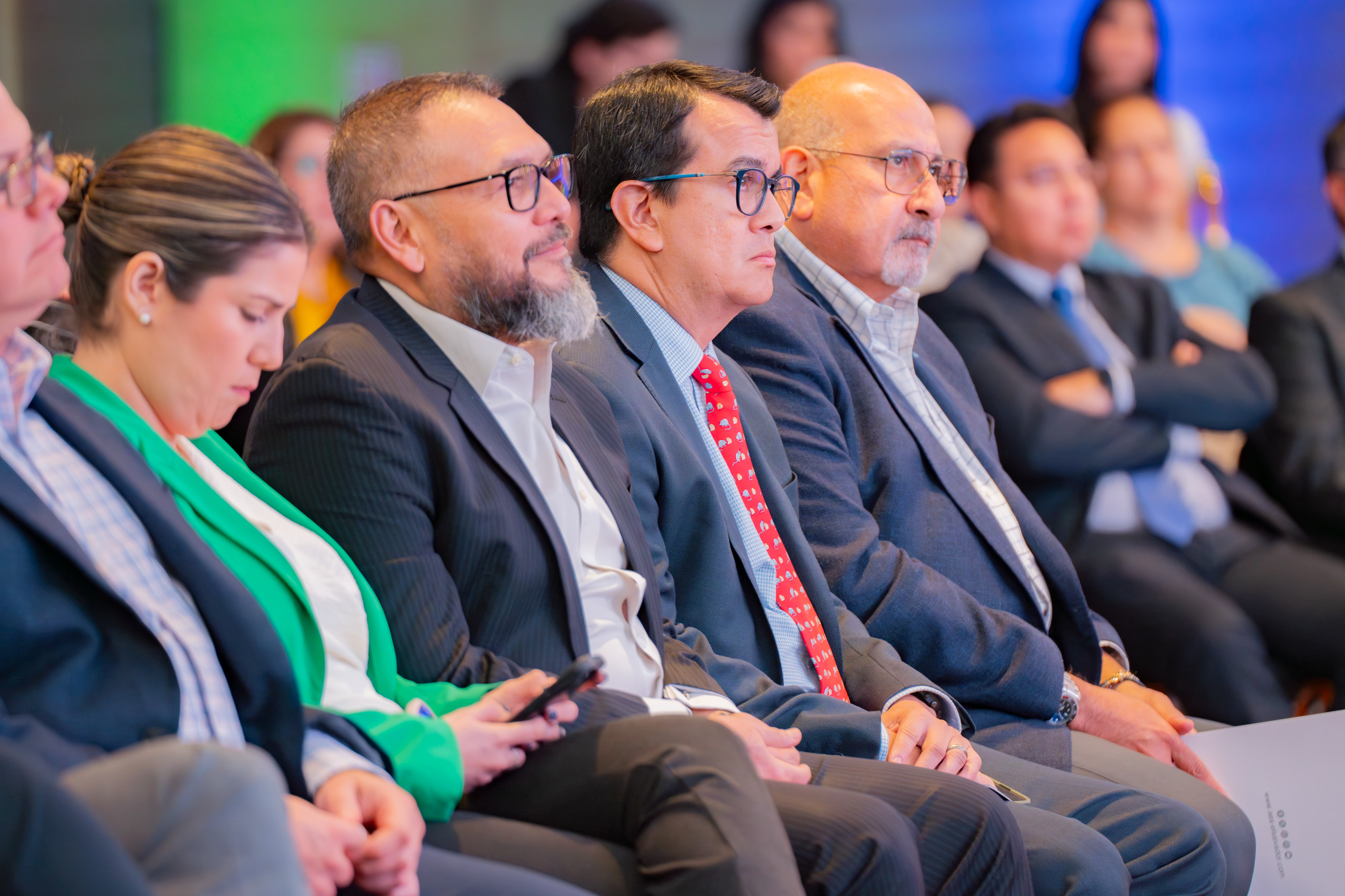 A group of business leaders and professionals sit in a conference setting, dressed in formal attire. The attendees include men in suits and ties, as well as a woman wearing a green blazer. They appear attentive and focused, with a few holding devices or documents. The blurred background suggests an indoor event with other participants, creating a professional and engaging atmosphere.