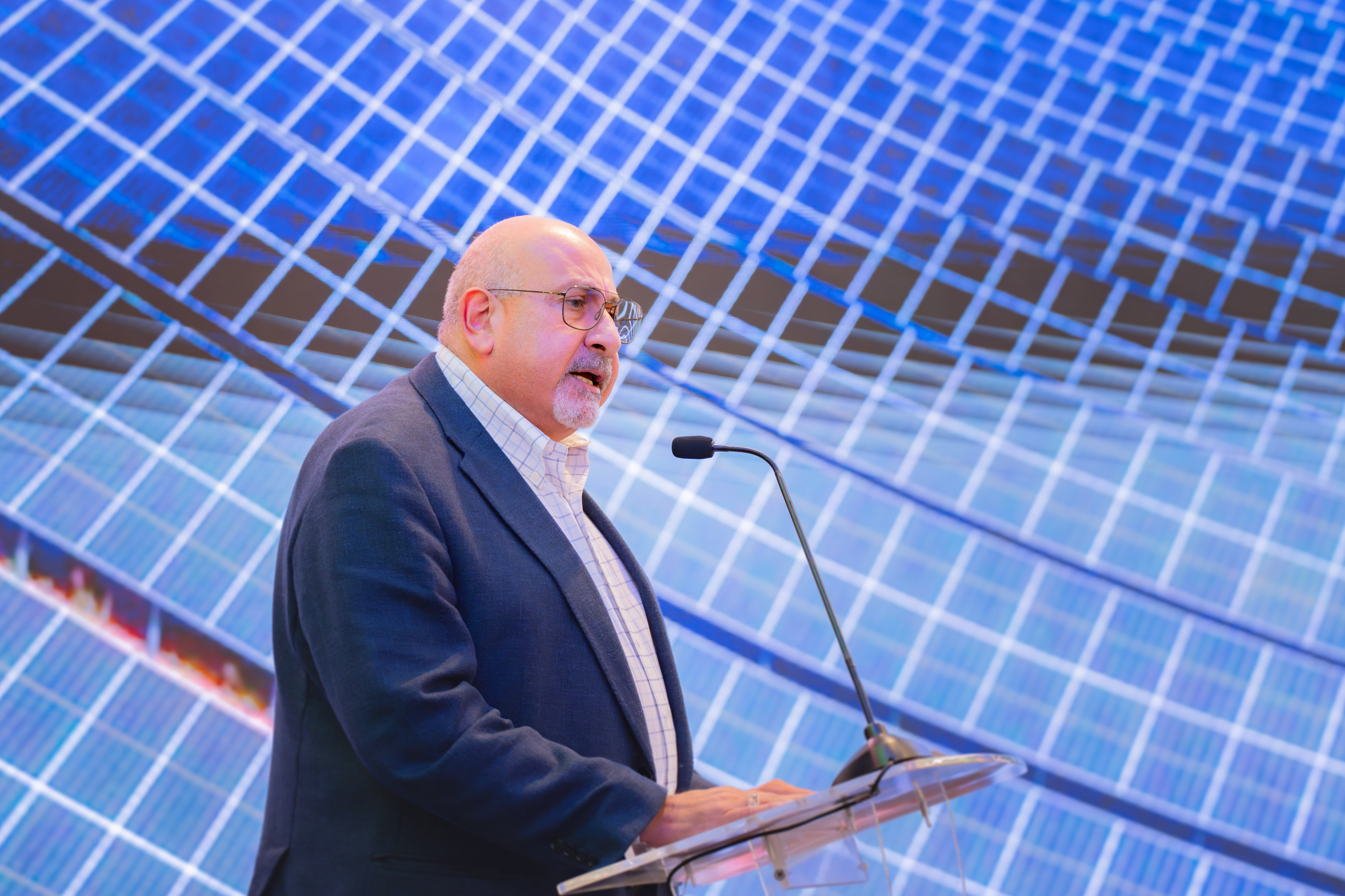 A speaker in a suit and glasses is presenting at a podium with a microphone. The background features a dynamic image of solar panels, creating a futuristic and innovative atmosphere. The speaker appears to be delivering a professional address, emphasizing topics related to renewable energy or sustainability.