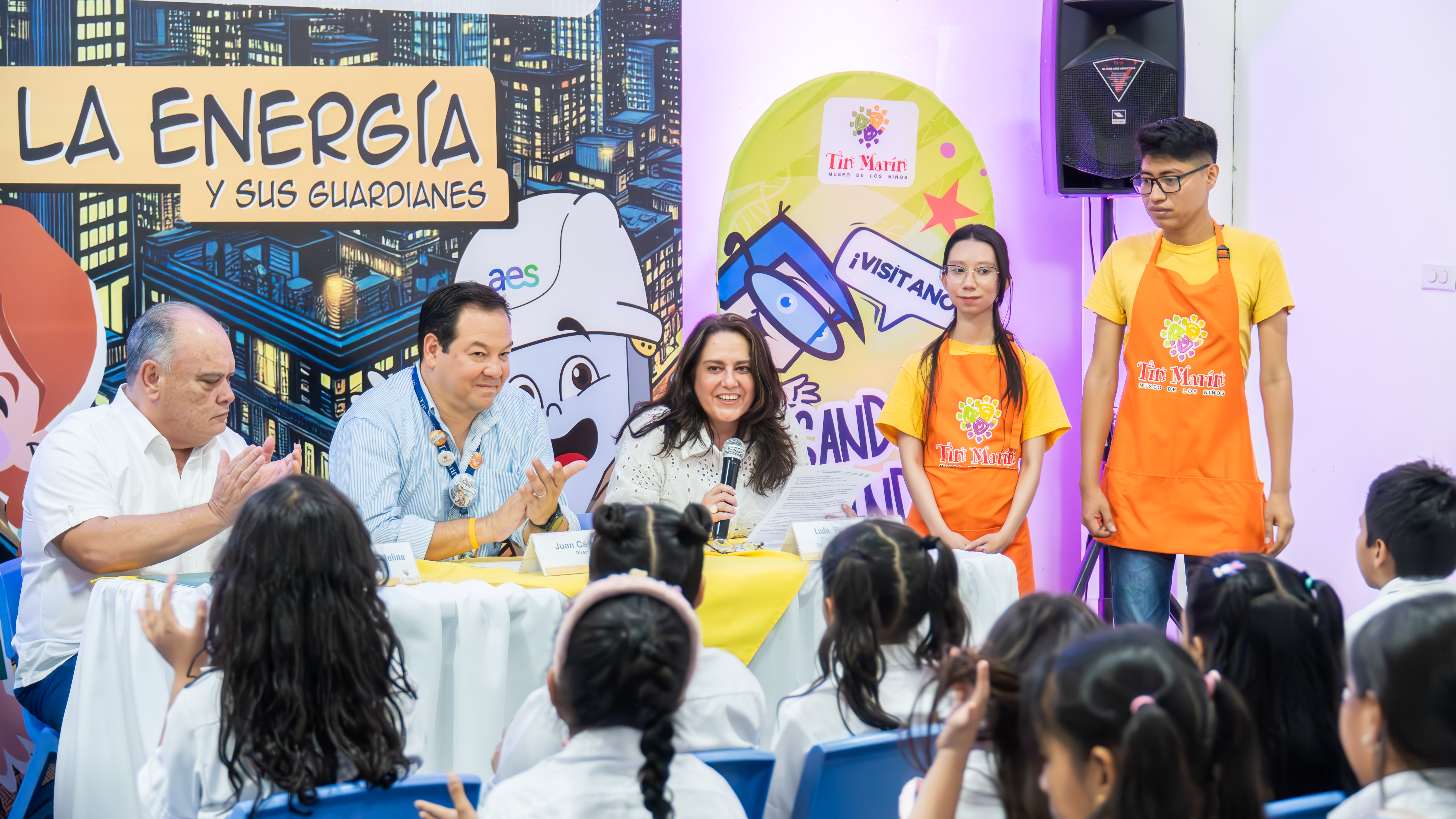 A panel of three adults sits at a table in front of a group of children, engaging in an educational event about energy. The backdrop features colorful graphics with the words "La Energía y sus Guardianes" and AES branding. Two staff members in bright orange "Tin Marín" aprons stand nearby. The panelists, including a woman speaking into a microphone, appear to be leading an interactive session with the children.