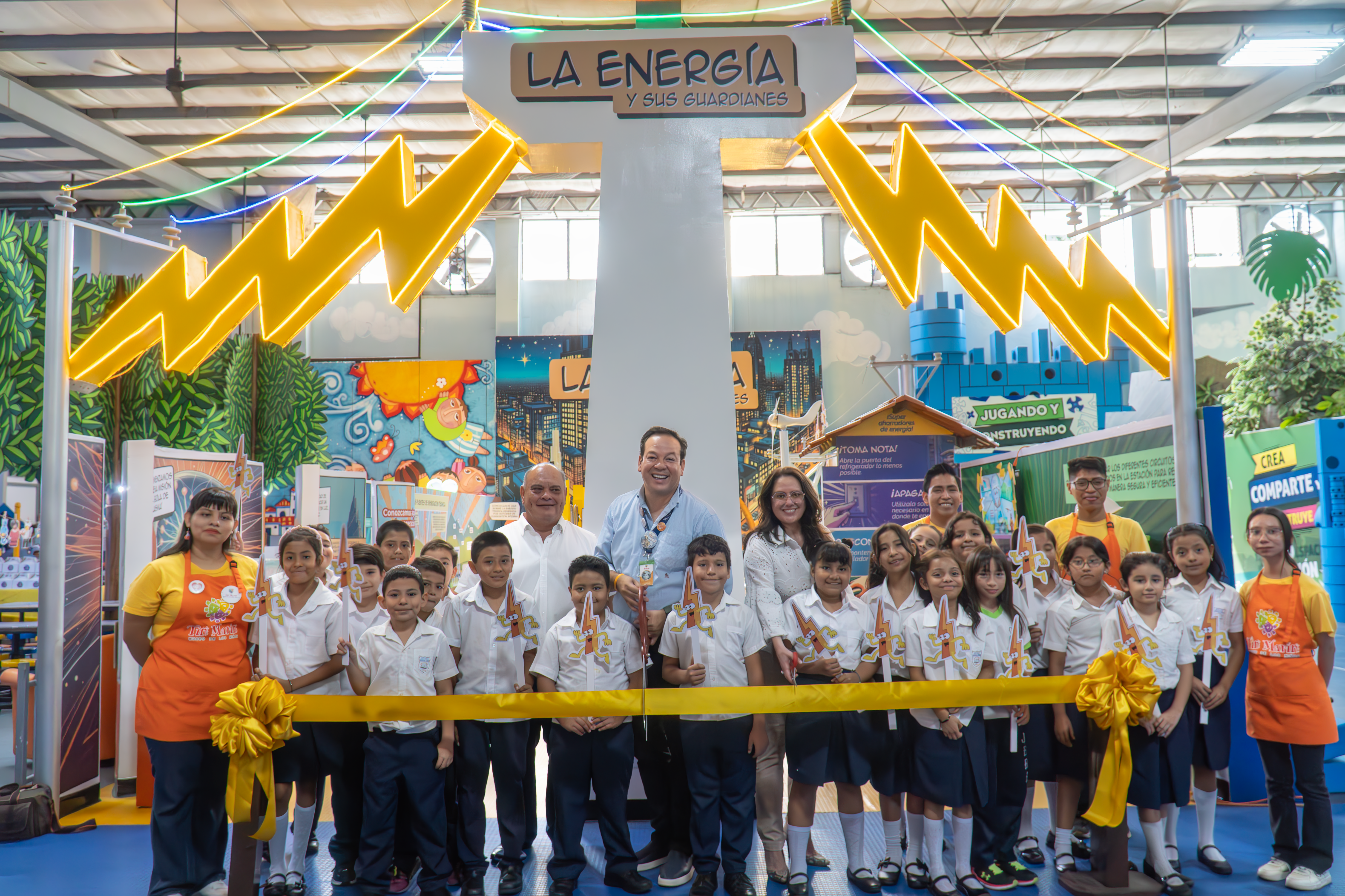 A group of school children in uniforms, along with AES representatives and staff in orange Tin Marín aprons, participate in a ribbon-cutting ceremony at an interactive energy exhibit. The exhibit features a large white pillar with the sign "La Energía y sus Guardianes" and bright yellow lightning decorations. The children hold lightning bolt-shaped cutouts, smiling for the camera in a colorful, educational space.