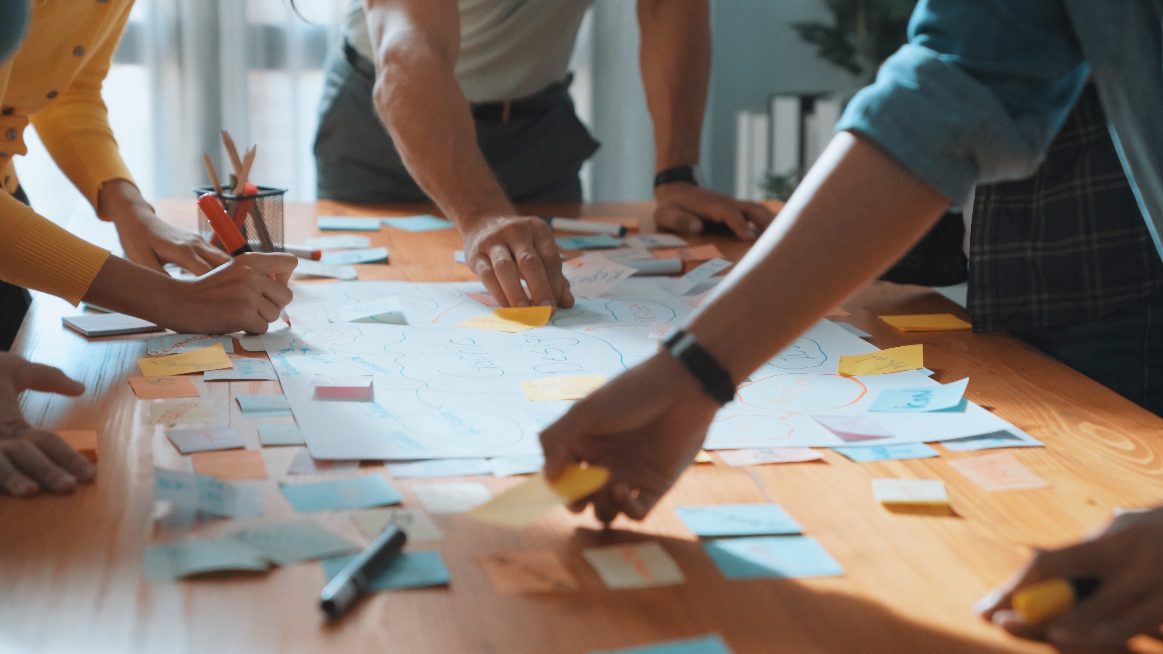 A group of people engaged in a brainstorming session around a wooden table, using colorful sticky notes and markers to write ideas on a large sheet of paper. Hands are actively placing notes and writing, while office supplies like pens and paper are scattered across the table. The background shows a well-lit workspace with natural light coming from windows.