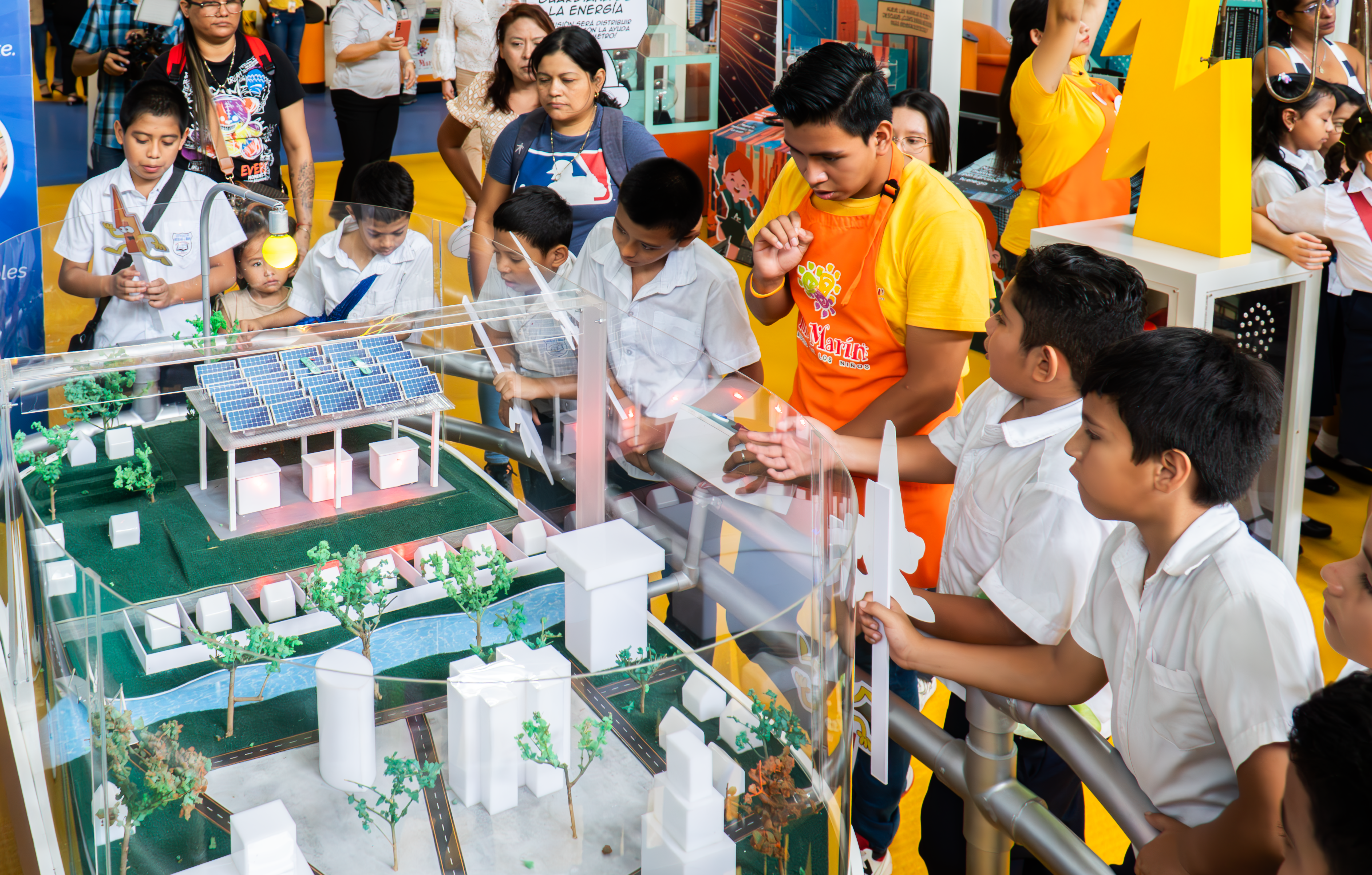 A group of school children in white uniforms gather around an interactive energy exhibit featuring a miniature model of a sustainable city with solar panels and wind turbines. A staff member in an orange Tin Marín apron explains the exhibit, while children observe and hold lightning bolt-shaped cutouts. The display is enclosed in glass, and the background features additional educational exhibits and attendees.