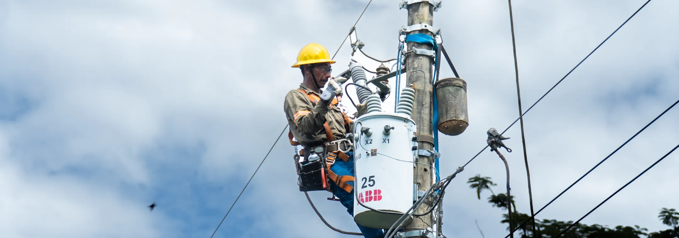 A utility worker performing maintenance on a utility pole against a bright blue sky with scattered white clouds. The worker, dressed in a brown uniform with reflective safety strips, wears a yellow hard hat, white gloves, and a safety harness. He is securely attached to the pole with climbing gear and tools strapped to his waist. The worker appears focused on his task, demonstrating professionalism and safety in electrical grid maintenance.