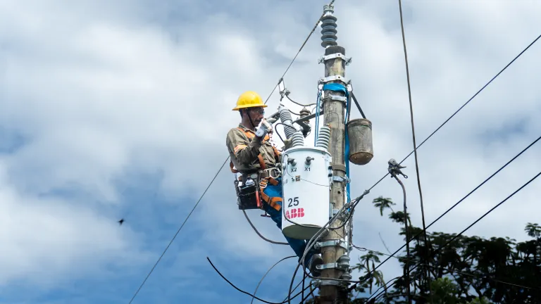 A utility worker performing maintenance on a utility pole against a bright blue sky with scattered white clouds. The worker, dressed in a brown uniform with reflective safety strips, wears a yellow hard hat, white gloves, and a safety harness. He is securely attached to the pole with climbing gear and tools strapped to his waist. The worker appears focused on his task, demonstrating professionalism and safety in electrical grid maintenance.