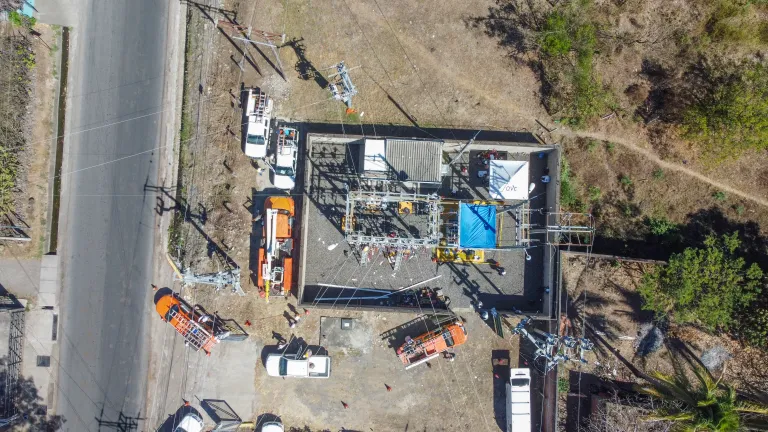 Aerial view of an electrical substation under maintenance, with workers in safety gear performing tasks. Several utility trucks, including orange and white vehicles, are parked nearby. A blue tent and a white tent with the "AES" logo are set up. Electrical lines extend from the substation, and a paved road runs alongside. Surrounding the area are patches of dry grass, trees, and a dirt path.