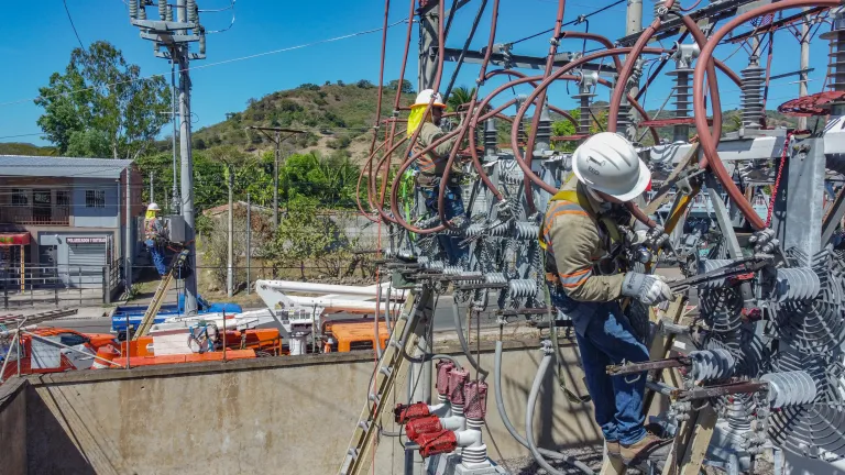 Two electrical workers in safety gear and helmets perform maintenance on a high-voltage substation. One worker is using tools to adjust equipment while standing on a ladder among thick red and black cables. Another worker is elevated on a nearby power pole. Several orange and white utility trucks are parked along the street, with a building and green hills visible in the background.