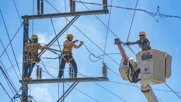 Three electrical workers in safety gear and helmets repair power lines on a transmission tower. Two workers are secured with harnesses on the metal structure, while a third is elevated in a bucket truck using insulated gloves to handle high-voltage wires. The background shows a bright blue sky with power lines stretching across the image.